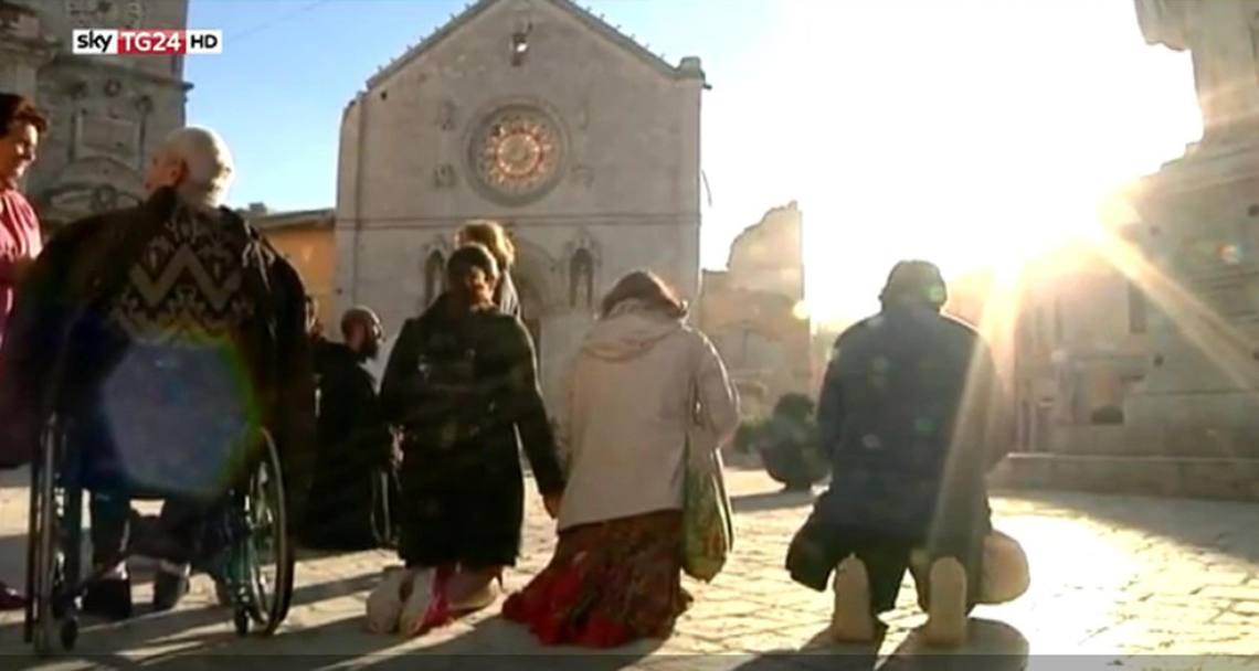 Fedeli inginocchiati in preghiera di fronte a quel che resta della cattedrale di San Benedetto. Ansa